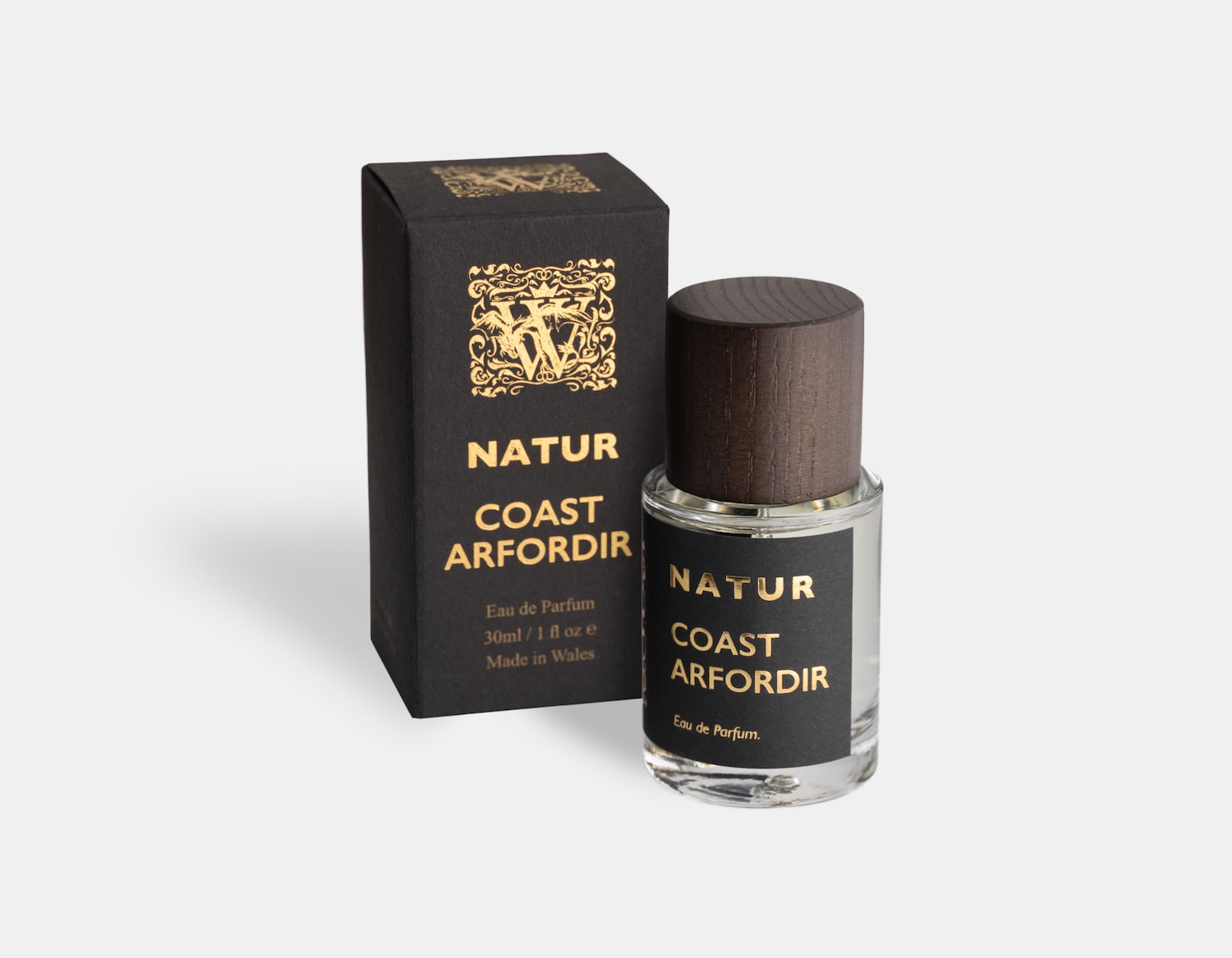 a cylindrical perfume bottle with a dark wood cap. The bottle has a black label with gold foil print, and is stood in front oF a matching black box with gold printing