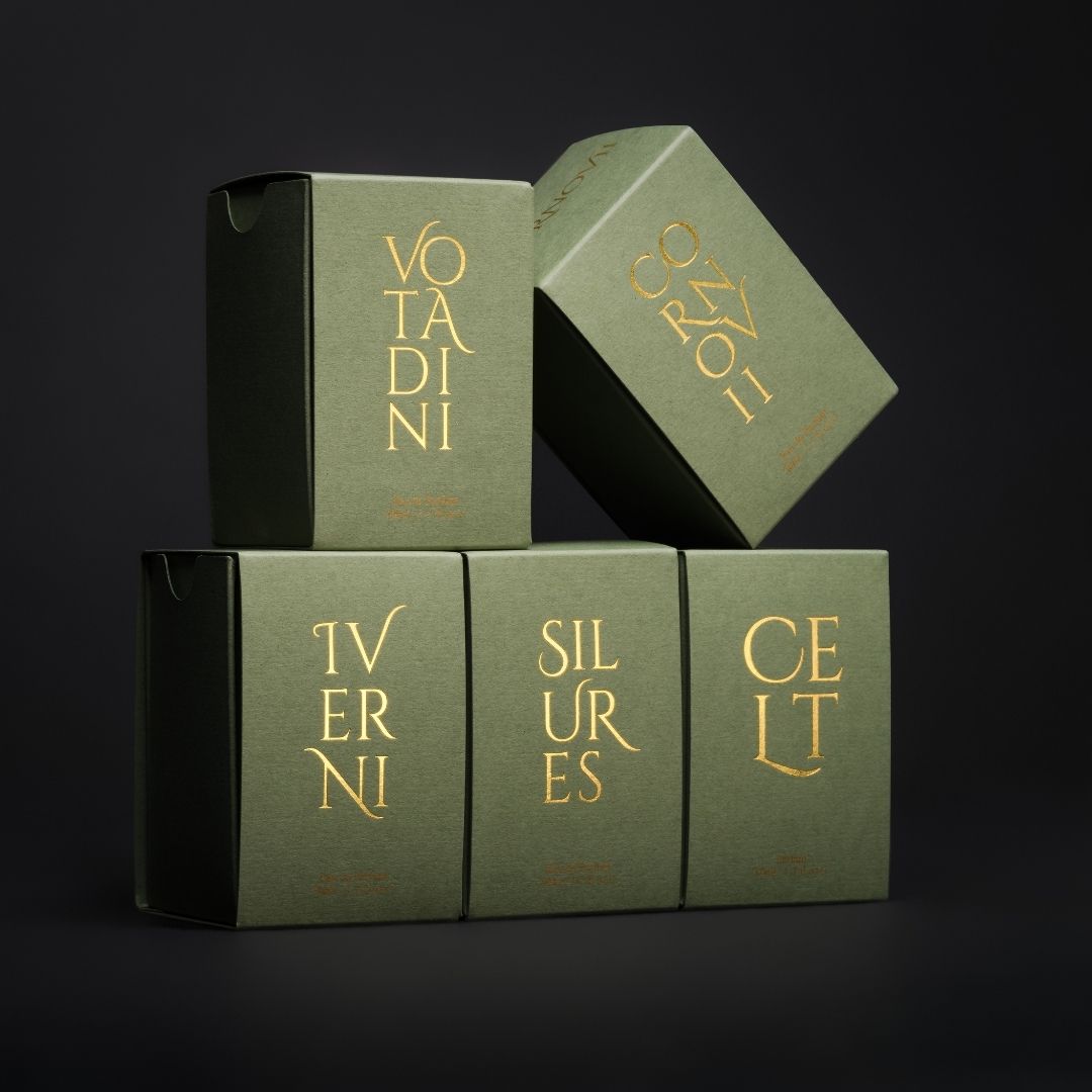 Five small square product boxes in deep sage green with gold foil lettering, stacked in a pyramid arrangement against a dark background. The boxes display names including Votadini, Iverni, Silures, Celt and Cornivii suggesting a collection of Wales Perfumery fragrances.