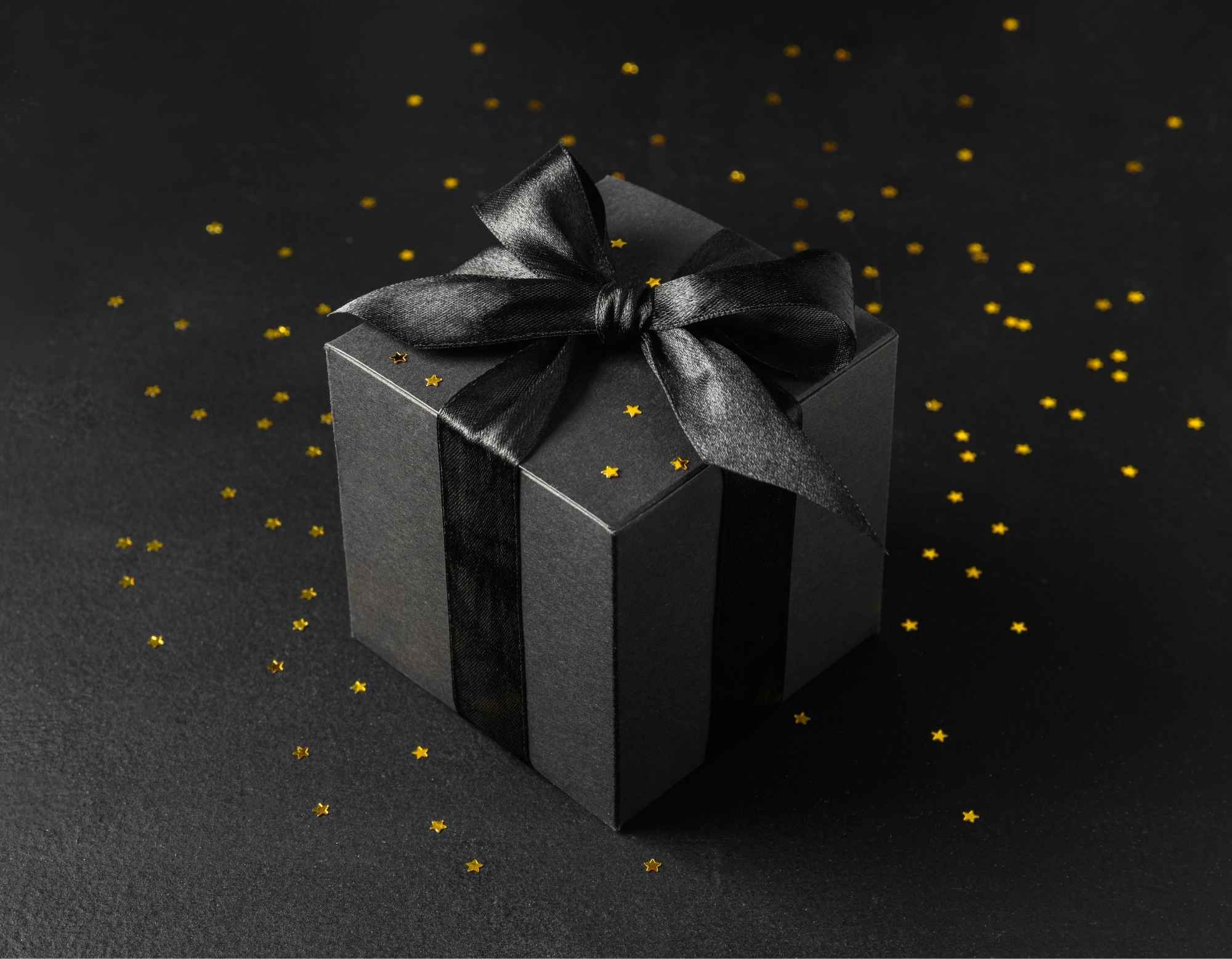 A black gift box tied with a black satin ribbon and bow, on a dark background scattered with small gold sparkles.