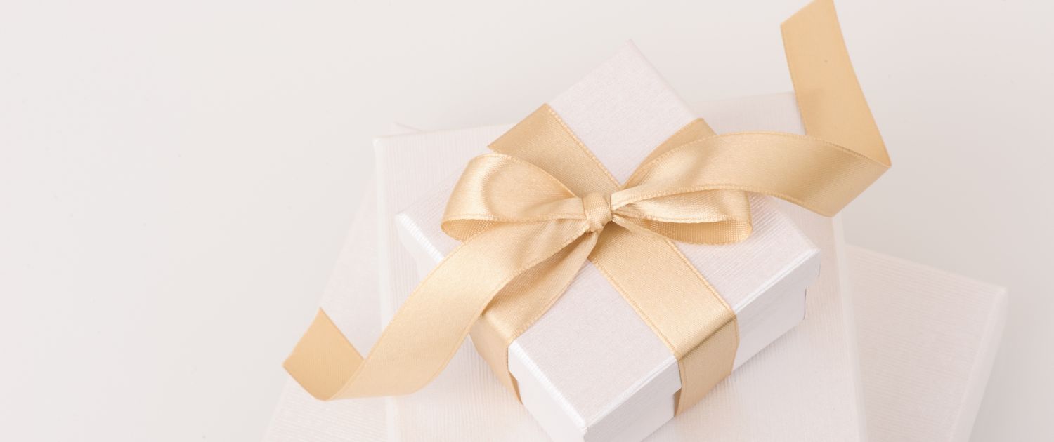 A white gift box tied with a gold satin ribbon and bow, resting on a soft cream surface.