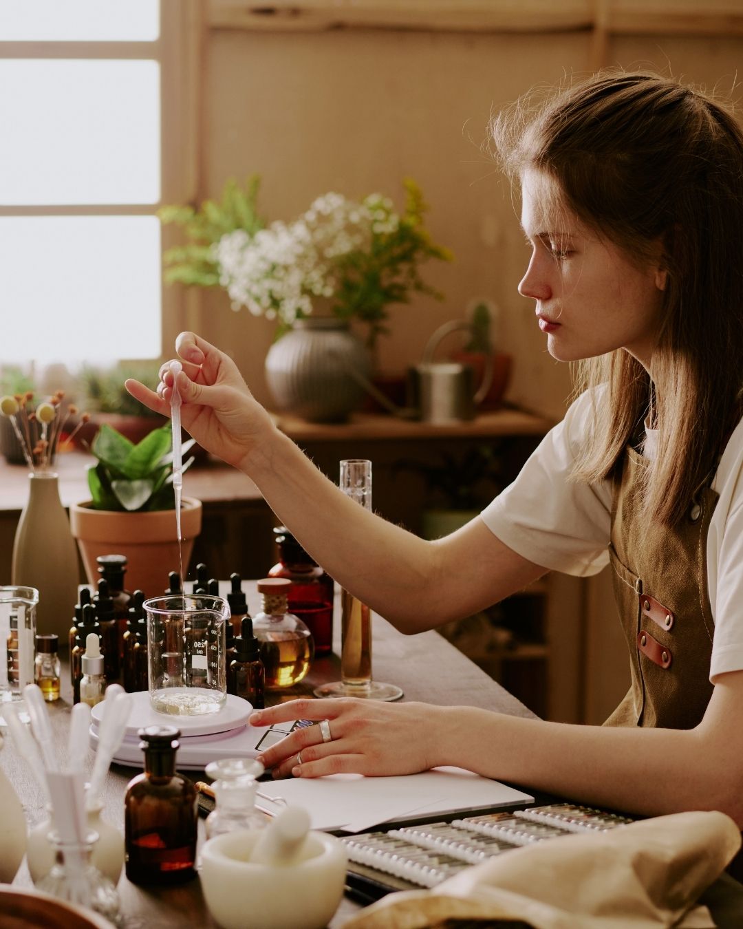 A perfumer working at a studio workbench, carefully measuring ingredients with a pipette, surrounded by amber glass bottles, a glass beaker on a small scale, and botanicals in the background.