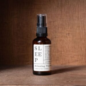 Amber glass spray bottle labelled "Sleep, Relaxing Pillow Mist" by Wales Perfumery, standing against a brown wooden backdrop.
