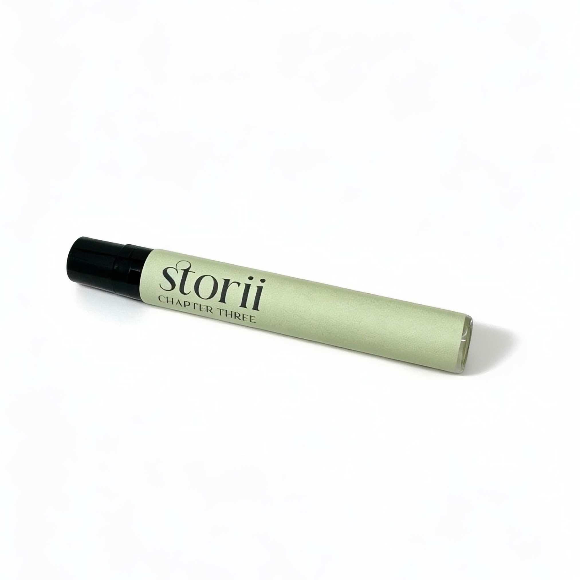 Storii Chapter Three 7ml perfume spray, sage green label with black cap, lying diagonally on a white background