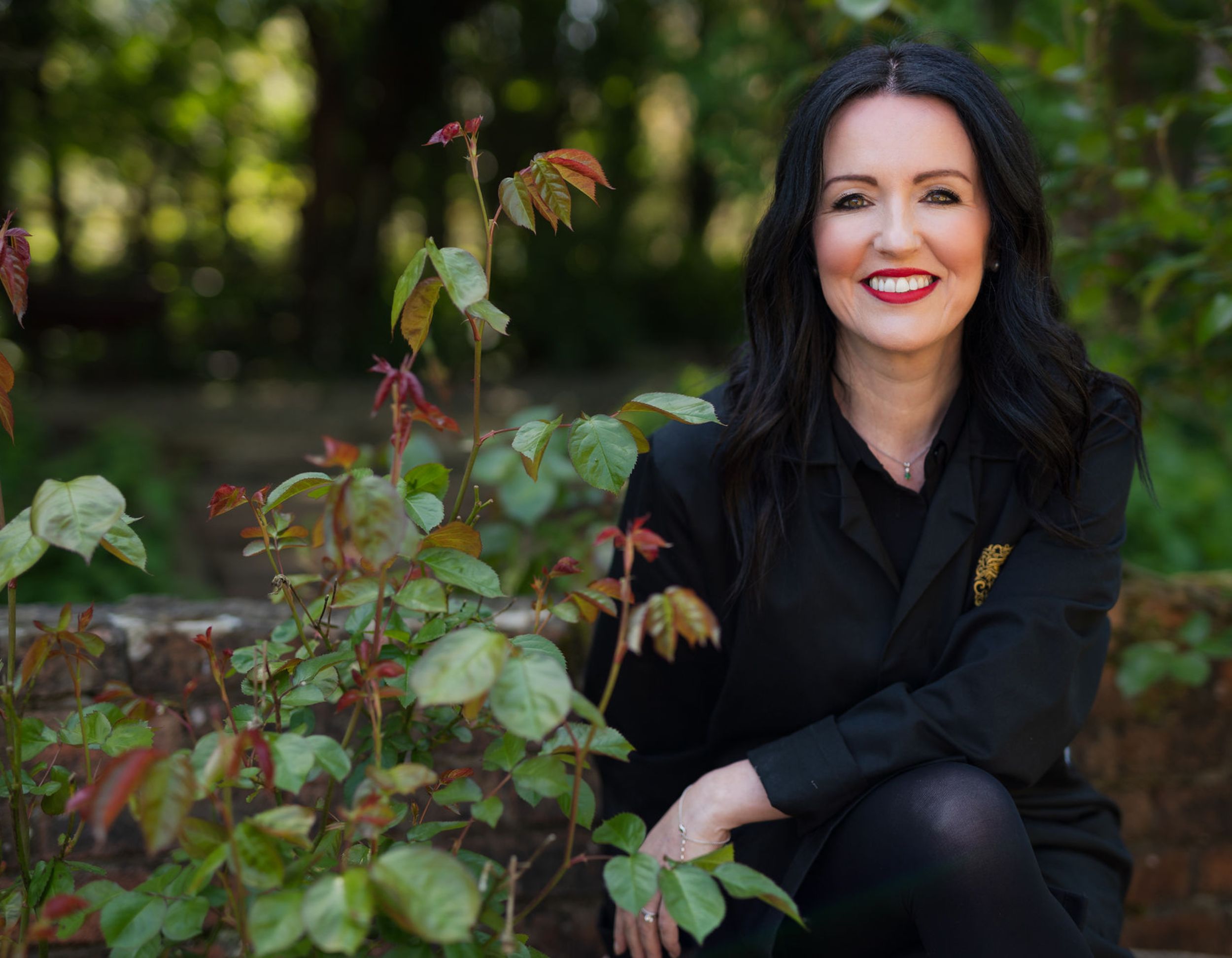 Louise Smith smiling outdoors beside a rose bush with autumn-toned leaves, dressed in her black Wales Perfumery workwear, with greenery and a low stone wall behind her.