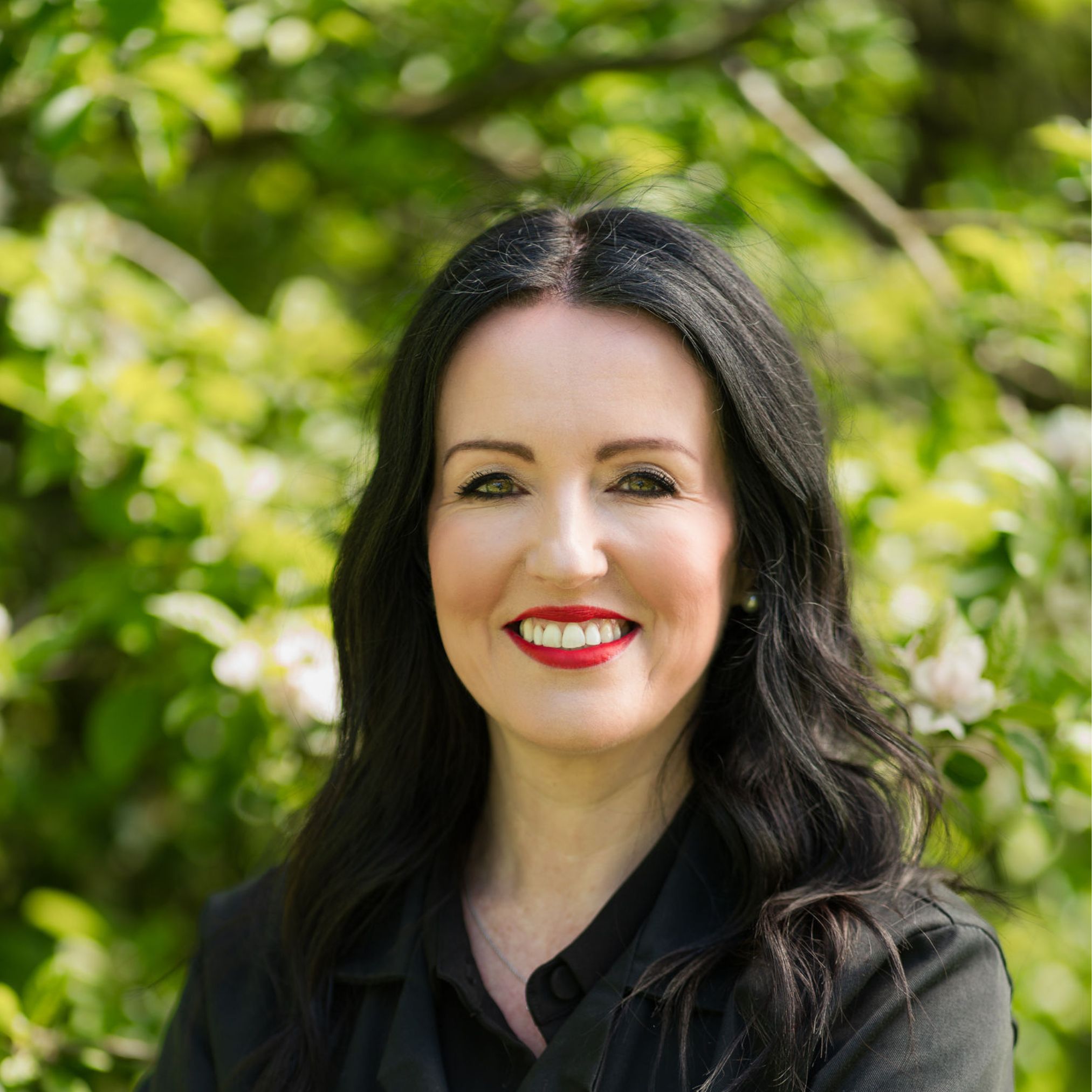 Louise Smith smiling outdoors in front of soft green foliage and blossom, wearing a black top, with her long dark hair loose.