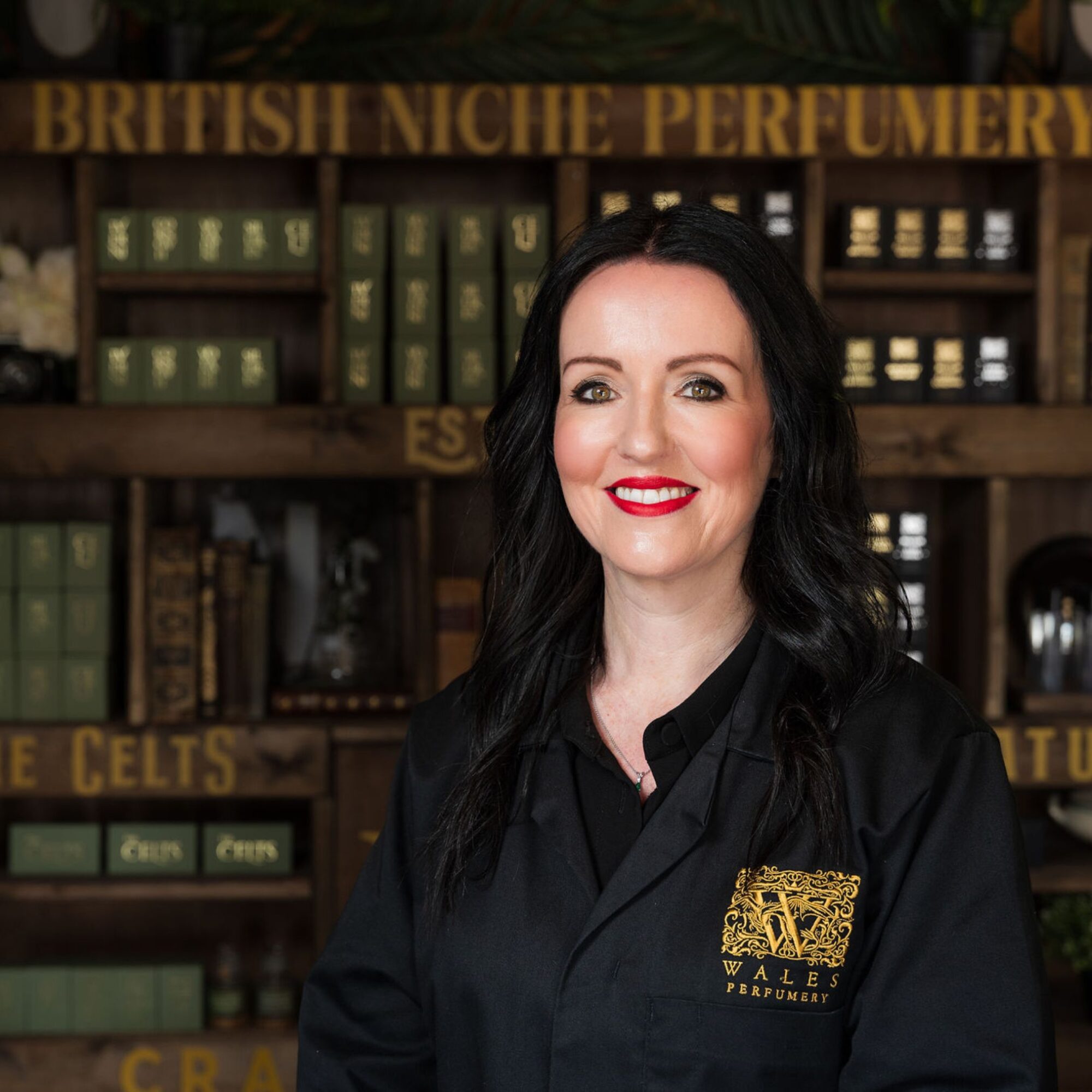 Louise Smith, founder of Wales Perfumery, smiling in a black perfumery coat with gold embroidered logo, standing in front of wooden shelving lined with books and fragrance boxes from the Celts and Natur collections, with 'British Niche Perfumery' lettering above.
