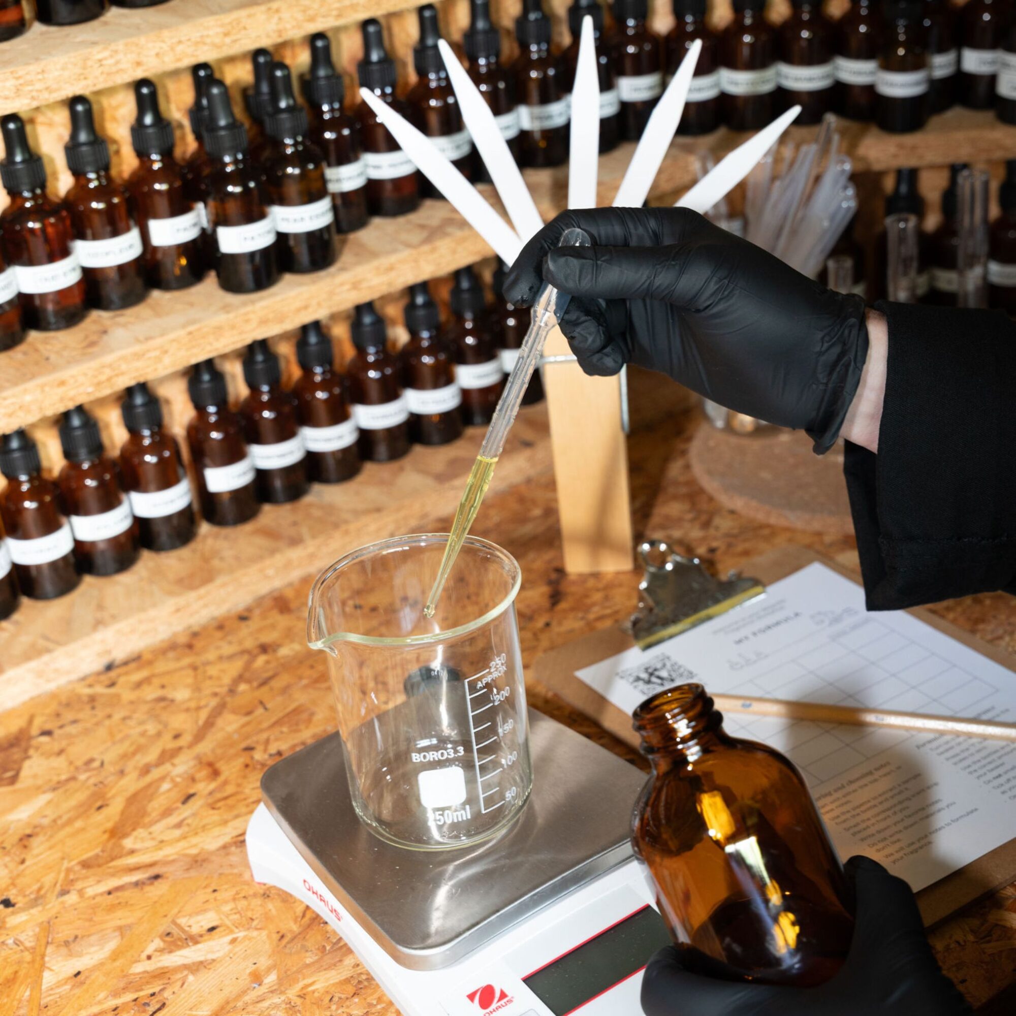 Gloved hand using a pipette to measure fragrance materials into a glass beaker on digital scales, with rows of amber glass bottles and blotting strips (scent testing strips) in the background, an amber bottle and handwritten formula sheet in the foreground.