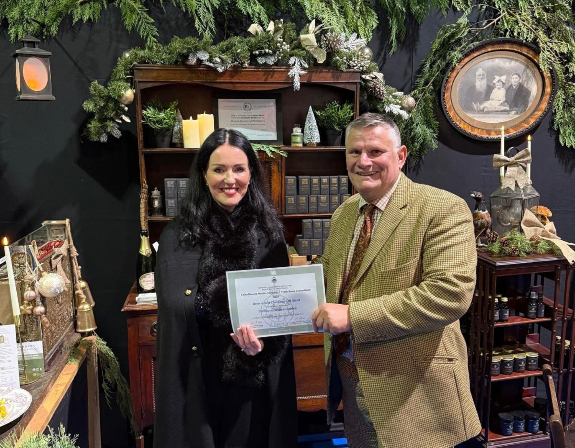 Louise of Wales Perfumery holding a Reserve Best Christmas Gift Stand certificate at the 2025 Royal Welsh Winter Fair, beside a man in a tweed jacket, in front of her festive stand with greenery, candles and shelves of perfume boxes.
